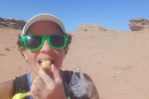 Woman with short black hair, wearing green sunglasses and white cap, stands in desert eating a cake.