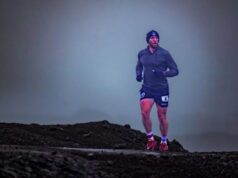 A male runners wearing shorts, long sleeve top and beanie hat runs across a remote stony landscape. The sky is very grey and dull.