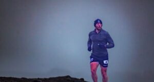 A male runners wearing shorts, long sleeve top and beanie hat runs across a remote stony landscape. The sky is very grey and dull.
