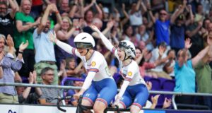 Two female cyclists on a tandem wave joyfully to a crowd in a cycling arena