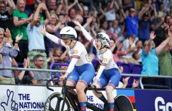 Two female cyclists on a tandem wave joyfully to a crowd in a cycling arena