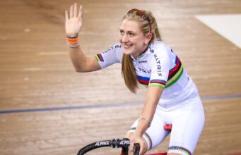 Laura Kenny in white cycling kit on her track bike waves to the crowd with a smile on her face