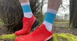 Very clean red trail shoes, on the feet on a runner, standing on a mossy rock in woodland, about to head off for a trail run.