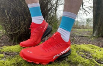 Very clean red trail shoes, on the feet on a runner, standing on a mossy rock in woodland, about to head off for a trail run.