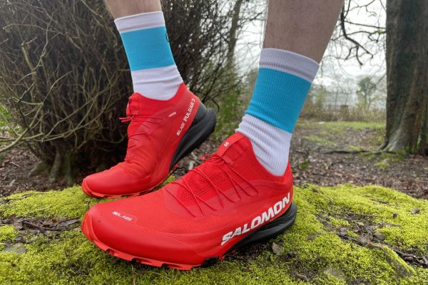 Very clean red trail shoes, on the feet on a runner, standing on a mossy rock in woodland, about to head off for a trail run.