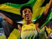 Jamaican woman with short cropped curly hair holds aloft a Jamaican flag. She is wearing a vest top in matching colours of yellow, green and black.