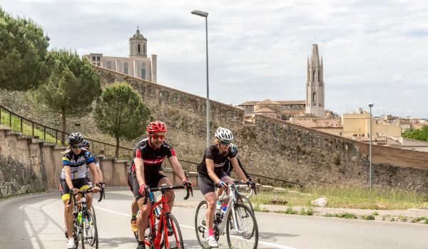 Three cyclists on a road somewhere hot and dusty with old stone buildings with towers behind them