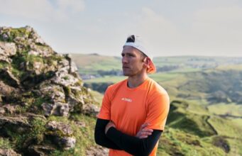 Male athlete wearing orange T-shirt over long sleeve black top, white cap, and orange bone conducting headphones, stands with arms folded and gazes off camera. The landscape behind him looks green and rocky.
