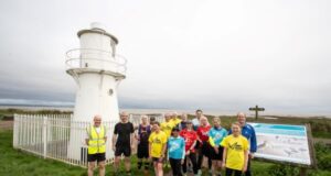 A group of runners stand in front of a small white lighthouse by the sea