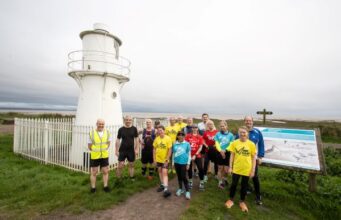 A group of runners stand in front of a small white lighthouse by the sea