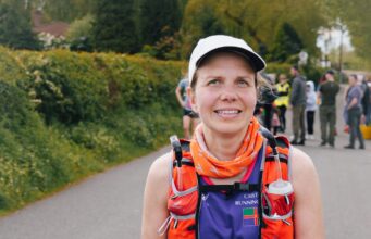 Lady in cap, running clothes and running hydration vest smiles at camera. She is wearing a race number, 438