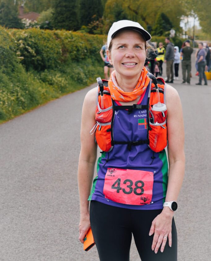 Lady in cap, running clothes and running hydration vest smiles at camera. She is wearing a race number, 438