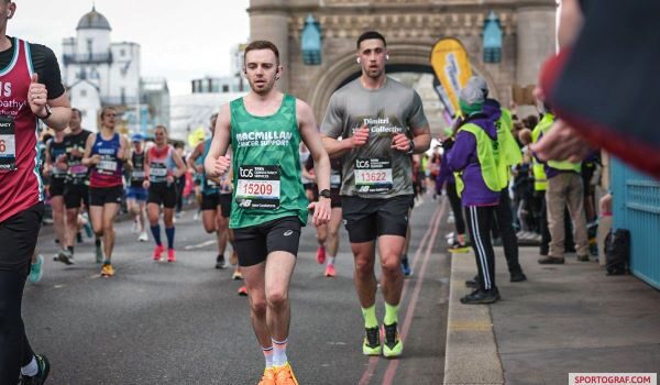 Young man in a Macmillan green vest, black shorts and orange trainers runs across Tower Bridge on the London Marathon