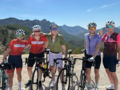 Five women each standing next to a bike smile for the camera in a sunny, mountainous landscape.