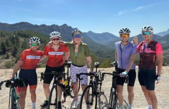 Five women each standing next to a bike smile for the camera in a sunny, mountainous landscape.