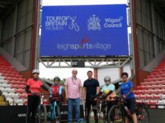 4 cyclists with bikes and two people without bikes stand underneath a huge sign reading Tour of Britain Women Leigh Sports Village