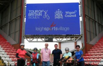4 cyclists with bikes and two people without bikes stand underneath a huge sign reading Tour of Britain Women Leigh Sports Village