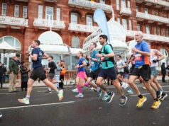 Runners run past red brick buildings at speed. One of them has a pace flag on his back.