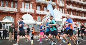 Runners run past red brick buildings at speed. One of them has a pace flag on his back.
