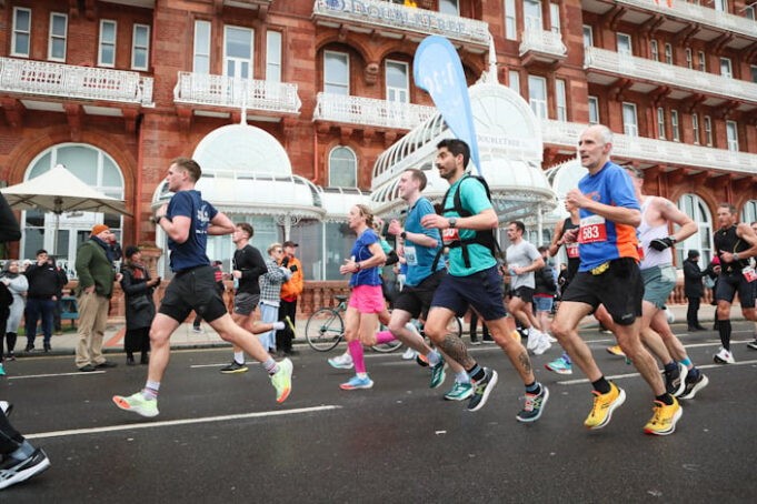 Runners run past red brick buildings at speed. One of them has a pace flag on his back.