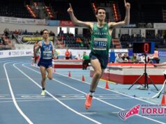 Man on running track in green vest holds arms in air in victory as he crosses finish line
