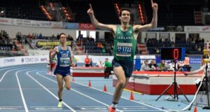 Man on running track in green vest holds arms in air in victory as he crosses finish line