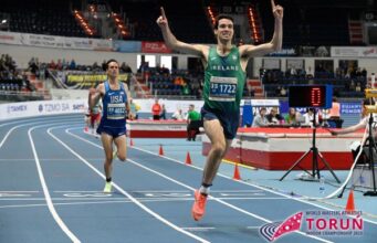 Man on running track in green vest holds arms in air in victory as he crosses finish line
