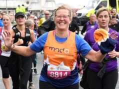 A running woman wearing glasses and an orange mnda charity vest smiles at the camera