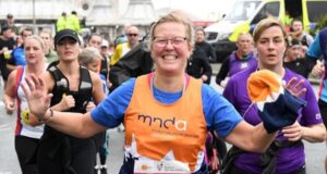 A running woman wearing glasses and an orange mnda charity vest smiles at the camera