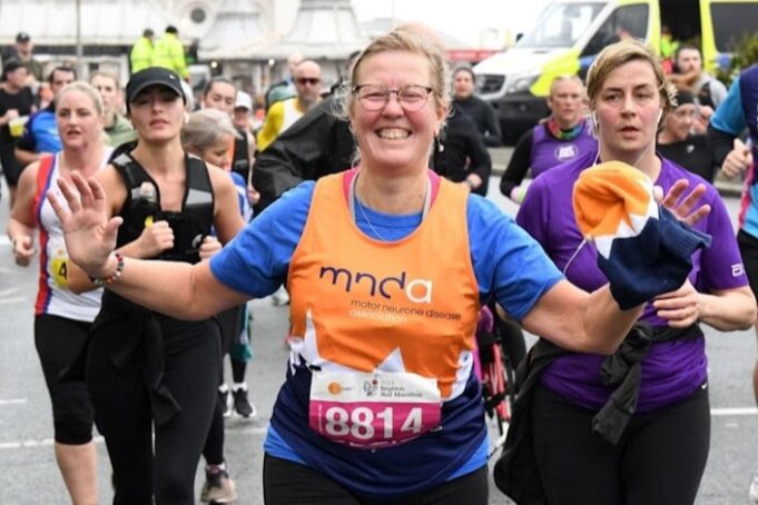 A running woman wearing glasses and an orange mnda charity vest smiles at the camera