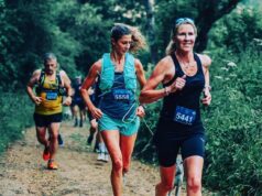 A woman in black vest and shorts runs along a forest trail with other runners, all wearing race numbers.