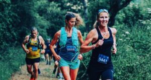 A woman in black vest and shorts runs along a forest trail with other runners, all wearing race numbers.