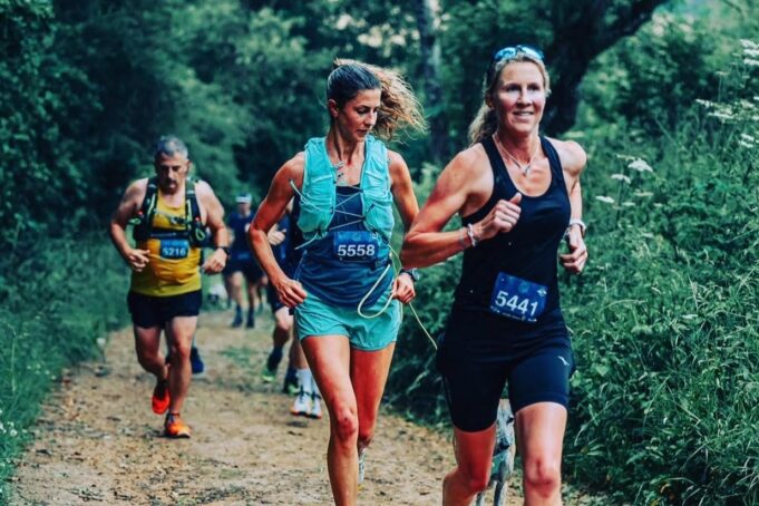A woman in black vest and shorts runs along a forest trail with other runners, all wearing race numbers.