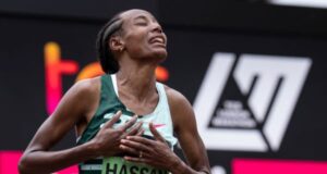 Woman clasps hands to heart in joy. She's wearing a running vest with HASSAN printed on her number.