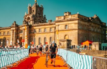 Triathletes run through a transition area with bikes in front of a spectacular palace