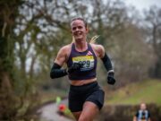 Female runner smiles, she's wearing a black crop vest, black shorts, black arm sleeves, and has her blonde hair in a pony tail