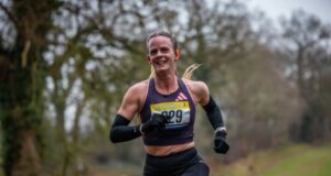 Female runner smiles, she's wearing a black crop vest, black shorts, black arm sleeves, and has her blonde hair in a pony tail
