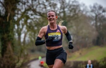 Female runner smiles, she's wearing a black crop vest, black shorts, black arm sleeves, and has her blonde hair in a pony tail