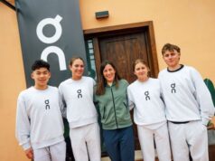4 young athletes in cream tracksuits stand with an older lady in green zip up top in front of an orange building with a sign saying On