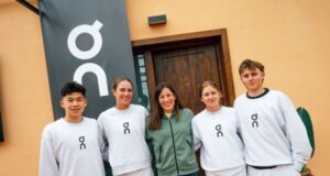 4 young athletes in cream tracksuits stand with an older lady in green zip up top in front of an orange building with a sign saying On