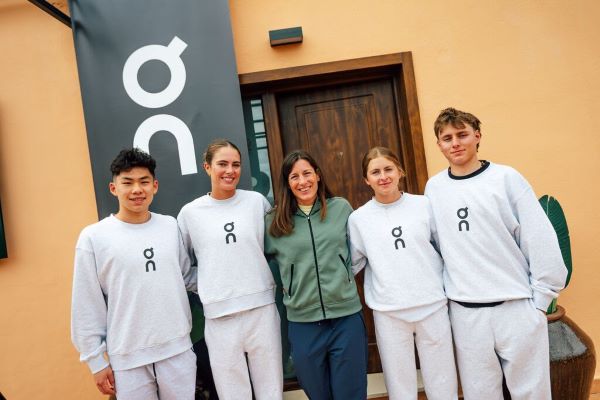 4 young athletes in cream tracksuits stand with an older lady in green zip up top in front of an orange building with a sign saying On