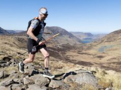 Male runner in shorts, T-shirt race vest is carrying poles while moving across a rocky mountain-top. He has a mohican, and is wearing a headband and dark sunglasses. He is smiling.