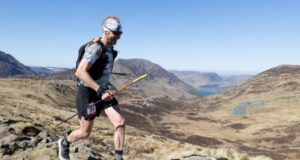 Male runner in shorts, T-shirt race vest is carrying poles while moving across a rocky mountain-top. He has a mohican, and is wearing a headband and dark sunglasses. He is smiling.
