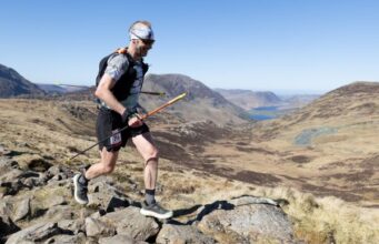 Male runner in shorts, T-shirt race vest is carrying poles while moving across a rocky mountain-top. He has a mohican, and is wearing a headband and dark sunglasses. He is smiling.