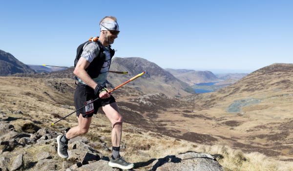 Male runner in shorts, T-shirt race vest is carrying poles while moving across a rocky mountain-top. He has a mohican, and is wearing a headband and dark sunglasses. He is smiling.