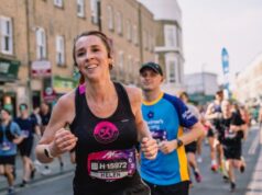 Female runner in black vest with long brown hair in plaits, runs amongst many other runners on an urban street.