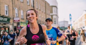 Female runner in black vest with long brown hair in plaits, runs amongst many other runners on an urban street.