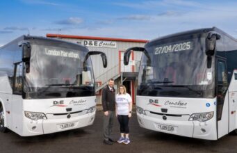 A man and a woman stand in front of two coaches at a depot