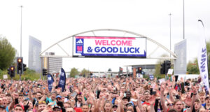 A large crowd of people cheer for the camera at the start line of the Manchester Marathon which reads Welcome and Good Luck