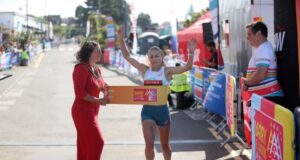 Woman runs through finish tape of race with arms held high, while another woman in a red dress holds the tape for her